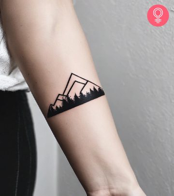 A line-art tattoo on the forearm of a mountain with a pine forest