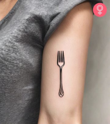 A woman wearing a minimalist fork tattoo on the upper arm.